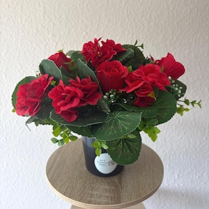 May include: A bouquet of vibrant red artificial roses and green foliage arranged in a black cylindrical pot. The container features a small white circular label on the front. The arrangement includes multiple blooming red flowers and round leaves.