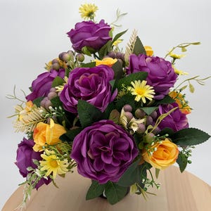 May include: A floral arrangement featuring deep purple roses and vibrant yellow flowers, accented with small green berries and green leaves. The bouquet includes various flower types, including small daisy-like blossoms and rose buds, creating a dense, colourful display of artificial botanical elements.