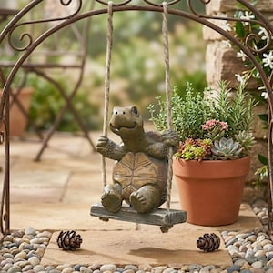 Op de afbeelding: Een bruin schildpadbeeldje zit op een kleine houten schommel die met twee touwen aan een gebogen metalen frame hangt. De schildpad heeft een textuur op het schild en staat naast een terracotta pot gevuld met diverse groene vetplanten.