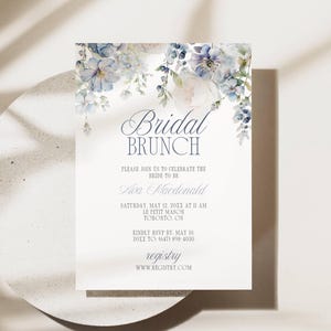 May include: A white invitation card with floral watercolor accents in shades of blue and purple. The text "Bridal Brunch" is displayed in elegant script, with additional details about the event, including the bride's name, date, time, and location.