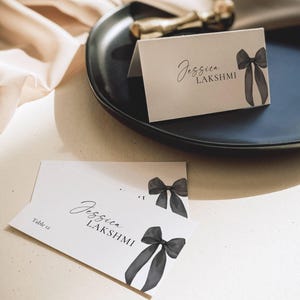 Könnte beinhalten: Elegante Tischkarten mit hellbeigem Hintergrund, mit den Namen "Jessica LAKSHMI" in einer stilvollen Schriftart. Jede Karte ist mit einer schwarzen Schleife im Aquarellstil verziert. Eine Karte liegt auf einem schwarzen Teller, eine andere auf einer weißen Oberfläche.