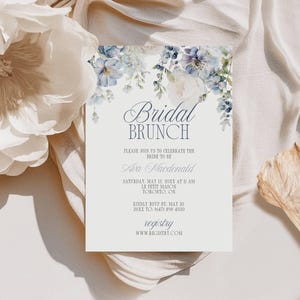 May include: A bridal brunch invitation with floral accents. The invitation features the text "Bridal Brunch" and details for the event, including the bride's name, date, time, and location. The design includes watercolor-style blue and white flowers.