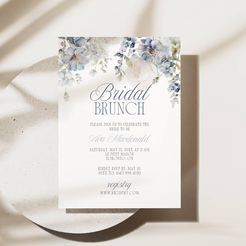 May include: A white invitation card with floral watercolor accents in shades of blue and purple. The text "Bridal Brunch" is displayed in elegant script, with additional details about the event, including the bride's name, date, time, and location.