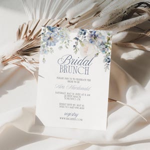 May include: A white invitation card with the text "Bridal Brunch" and floral accents in shades of blue and white. The card includes details for a bridal brunch, including the bride's name, date, time, and location.