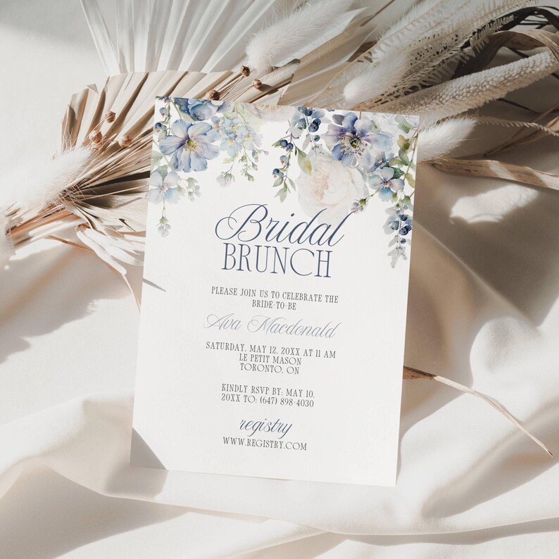 May include: A white invitation card with the text "Bridal Brunch" and floral accents in shades of blue and white. The card includes details for a bridal brunch, including the bride's name, date, time, and location.