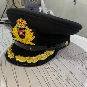 May include: A black peaked cap featuring a prominent gold embroidered emblem on the front, which includes a crown and a central flag design. The visor is adorned with gold leaf embroidery, and a black chin strap is secured by gold-toned buttons.