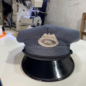 Op de afbeelding: Een donkerblauwe uniformpet met een zwarte lakleren klep en een zwarte gevlochten koordafwerking. Centraal op de voorzijde zit een metalen embleem met een adelaar, de tekst U.S. POST OFFICE, het nummer 1401 en LETTER CARRIER.