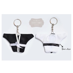 Peut inclure: Deux porte-clés miniatures en forme de kimono de karaté, un noir et un blanc, chacun avec un porte-clés argenté. Le kimono noir a un nœud blanc et le kimono blanc a une ceinture noire. Fond blanc.