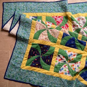 May include: A patchwork quilt featuring a repeating pinwheel pattern with green fabric blades. The quilt includes various floral, butterfly, and polka dot prints in blue, yellow, pink, and white, framed by a yellow border and a blue floral edge.