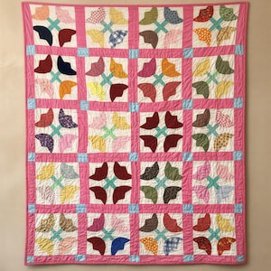 Puede incluir: Un edredón hecho a mano con un borde rosa y un patrón floral repetitivo. El edredón presenta cuadrados con cuatro diseños en forma de pétalos en varios colores, incluyendo rojo, azul, amarillo y verde, sobre un fondo blanco.