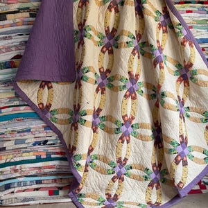 Op de afbeelding: Een quilt met een dubbel trouwringpatroon op een crèmekleurige achtergrond, met in elkaar grijpende ringen in diverse bloem- en geometrische patronen in geel-, groen- en roodtinten, afgewerkt met een effen paarse rand en paarse achterkant.