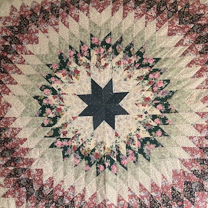 May include: A quilt featuring a central dark navy blue star surrounded by concentric layers of fabric patches. The design incorporates floral patterns in shades of pink, green, cream, and deep red, arranged in a geometric starburst pattern.
