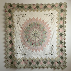 May include: A square quilt with a central floral design in pink and green, set against a white backdrop. It has a scalloped edge and a border of alternating green and pink squares, with leaf accents.