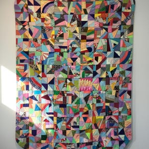 Può includere: Una colorata trapunta patchwork con una varietà di forme e motivi geometrici. La trapunta presenta quadrati, triangoli e altre forme in un mix di colori vivaci, tra cui rosa, blu, giallo e verde. La trapunta è appesa a un muro.