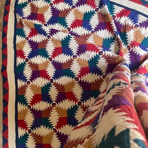 May include: A colorful quilt with a geometric pattern. The quilt features a repeating design of squares in various colors, including red, blue, green, purple, and tan. The quilt has a white border with a red and blue triangle trim.