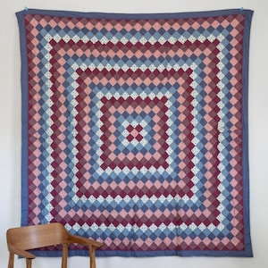 Könnte beinhalten: Eine große, quadratische Steppdecke mit einem sich wiederholenden Rautenmuster in Blau-, Rosa- und Burgundertönen. Die Steppdecke hängt an einer weißen Wand. Ein Holzstuhl befindet sich im Vordergrund. Die Steppdecke hat einen blauen Rand.