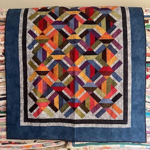May include: A quilt featuring a geometric pattern of multicolored fabric strips in shades of red, orange, yellow, green, blue, and purple. The central design is framed by a thin black border, a gray floral print border, and a wide solid blue border.