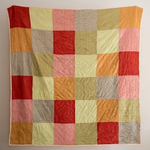 May include: A colourful patchwork quilt featuring a grid of squares in red, orange, pink, yellow, and grey. The quilt is displayed against a white wall, highlighting its handmade construction and vibrant design.