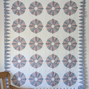May include: A large, handmade quilt with a white background and repeating fan-shaped patterns in shades of pink, blue, and floral prints. The quilt has a scalloped edge and a border with a floral pattern. The quilt is hanging on a white wall.