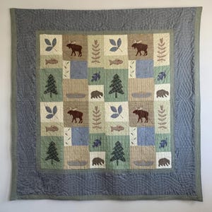 May include: A quilted throw blanket with a nature-themed design. The square pattern features images of moose, fish, trees, leaves, and bears in shades of brown, green, and blue. The blanket has a grey border.