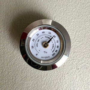 Puede incluir: Un higrómetro de color plateado con una cara blanca y una aguja negra. La cara tiene marcas para los niveles de humedad del 0 al 100 por ciento, con la aguja apuntando al 50 por ciento. Las palabras "VERY DRY" y "NORMAL" están impresas en la cara, junto con el texto "HYGRO 001".