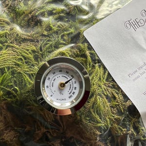 Könnte beinhalten: Ein rundes, silberfarbenes analoges Hygrometer mit weißem Zifferblatt, schwarzen Markierungen und einem Zeiger, der auf den Normalbereich zeigt. Das Gerät liegt auf grünem Moos in einem durchsichtigen Plastikbeutel neben einer weißen Karte.