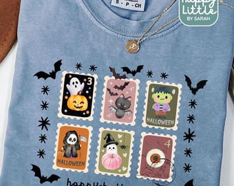 May include: Light blue t-shirt with a Halloween-themed design. The design features postage stamp-style graphics with a ghost, black cat, Frankenstein, Grim Reaper, and other spooky elements. The words "happy halloween" are printed at the bottom.