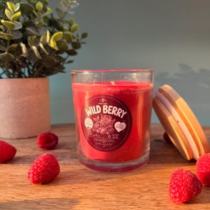 Könnte beinhalten: Eine rote Duftkerze in einem klaren Glasgefäß mit einem Holzdeckel. Das runde Etikett trägt die Aufschrift Wild Berry und zeigt Beerenillustrationen. Die Kerze ist von frischen Himbeeren auf einer Holzoberfläche umgeben.