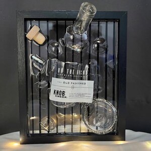 May include: A framed artwork featuring a clear glass bottle and a glass with the text "KNOB CREEK" and "THE OLD FASHIONED". The artwork includes a wooden cork and is set against a black background.