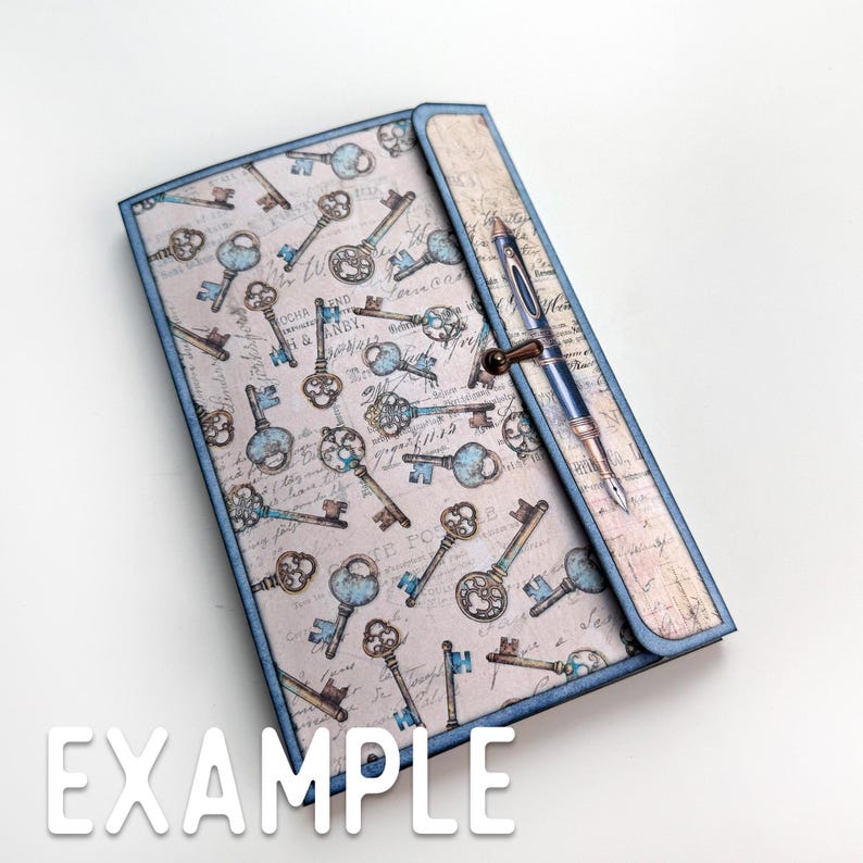 May include: A blue and beige journal with a decorative key pattern and a pen tucked into the cover. The pen has a gold clip and a black nib.