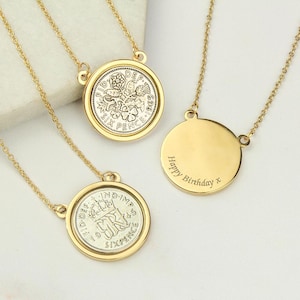 May include: Gold necklaces featuring coin pendants. The coins are set in gold frames and display detailed designs. One necklace has a blank, round pendant with "Happy Birthday x" engraved. The necklaces are presented on a white surface.
