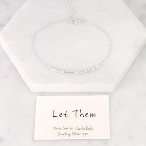 May include: A delicate silver bracelet with a Morse code design. The bracelet is displayed on a white marble surface, with a small card that reads "Let Them". The card also includes the text "Morse Code by: Charlie Boots Sterling Silver 925".