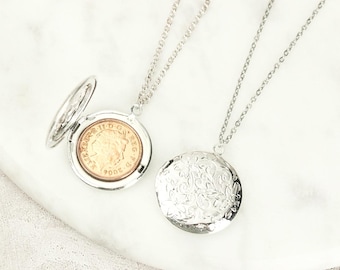 May include: Two silver-toned metal locket necklaces with thin chains. One locket is open, revealing a gold-toned 2004 British coin inside. The other locket is closed and features an intricate, embossed floral pattern on its circular surface.
