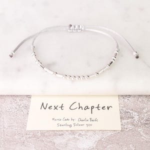 May include: A silver Morse code bracelet with a sliding knot closure. The bracelet is on a white surface with a card that reads "Next Chapter". The card also states "Morse Code by: Charlie Boots Sterling Silver 925".