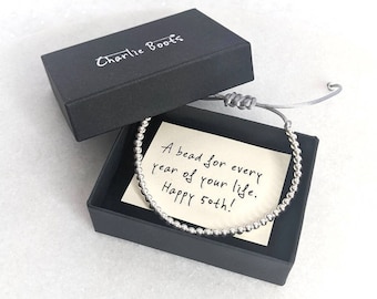 May include: A silver bead bracelet with a gray adjustable cord, presented in a black box. The box lid is open, revealing a handwritten note that reads, "A bead for every year of your life. Happy 50th!" The box is branded with "Charlie Boots".