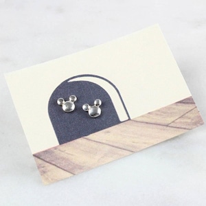May include: A pair of silver Mickey Mouse head stud earrings on a white card with a black and white illustration of an archway.