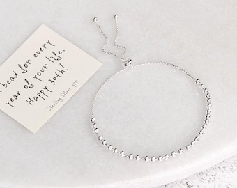 May include: A silver adjustable bracelet with small, round beads. A small card next to the bracelet reads "A bead for every year of your life. Happy 30th! Sterling Silver 925."