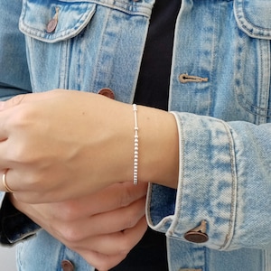 Op de afbeelding: Een delicate zilveren armband met een reeks kleine, gelijkmatig verdeelde kralen. De armband wordt om een pols gedragen, met een spijkerjasje op de achtergrond. Het ontwerp van de armband is eenvoudig en elegant, geschikt voor dagelijks gebruik.