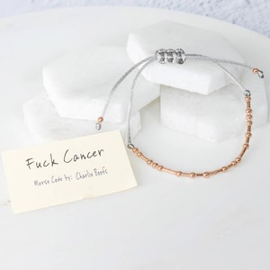 K&ouml;nnte beinhalten: Ein silbernes und ros&eacute;goldenes Armband mit Morsecode-Design. Das Armband liegt auf einer wei&szlig;en Oberfl&auml;che mit einer kleinen Karte mit der Aufschrift "Fuck Cancer" und "Morse Code by: Charlie Boots."