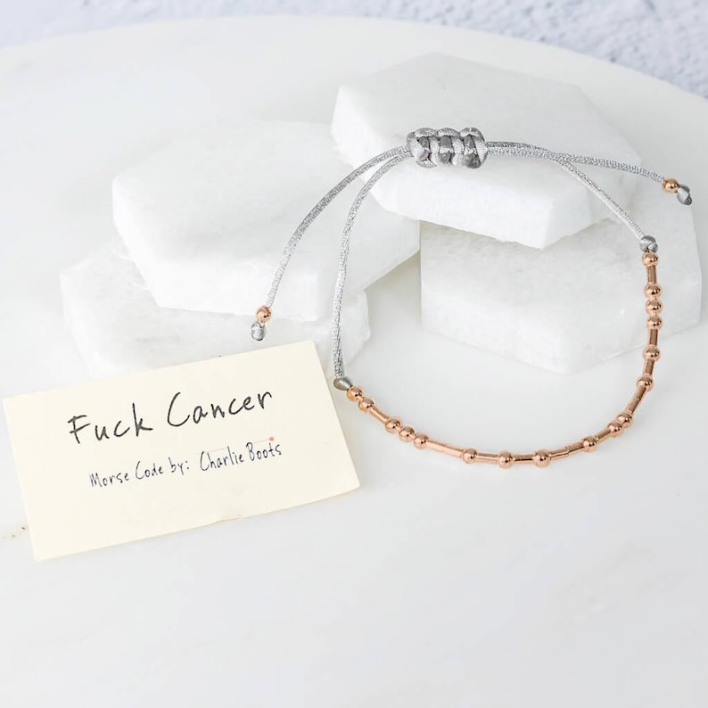 K&ouml;nnte beinhalten: Ein silbernes und ros&eacute;goldenes Armband mit Morsecode-Design. Das Armband liegt auf einer wei&szlig;en Oberfl&auml;che mit einer kleinen Karte mit der Aufschrift "Fuck Cancer" und "Morse Code by: Charlie Boots."