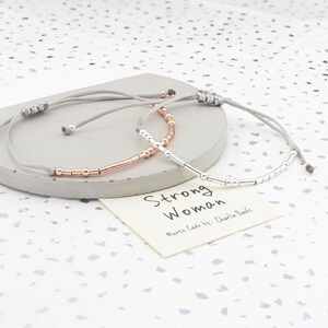 May include: Two adjustable bracelets with gray cords and silver and rose gold beads. The bracelets spell out "Strong Woman" in Morse code. The card reads "Morse Code by: Charlie Boots".