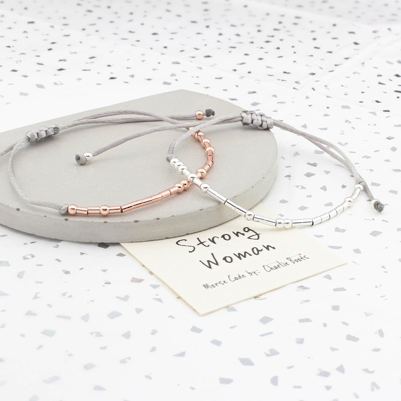 May include: Two adjustable bracelets with gray cords and silver and rose gold beads. The bracelets spell out "Strong Woman" in Morse code. The card reads "Morse Code by: Charlie Boots".