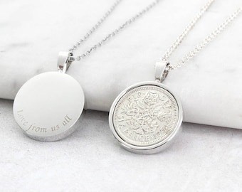 May include: Two silver pendant necklaces. One pendant is a blank, round disc with the engraved text "Love from us all". The other pendant features a coin design. Both necklaces have delicate silver chains and are displayed on a white surface.