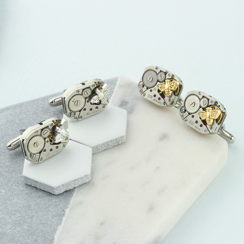 May include: A set of four silver cufflinks with a gold bee design. Each cufflink features a detailed clockwork design with a small ruby gemstone.