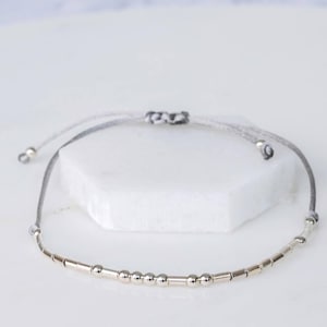 Puede incluir: Un brazalete delicado con una cadena plateada y peque&ntilde;as cuentas plateadas en un cord&oacute;n gris.
