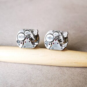 May include: A pair of silver cufflinks with a detailed clockwork design and a small silver butterfly on each. The cufflinks have a red jewel accent.