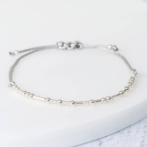 K&ouml;nnte beinhalten: Ein silbernes Perlenarmband mit einem verstellbaren grauen Band. Das Armband hat eine Reihe kleiner, runder Perlen und zylindrischer St&auml;be, die zu einem zarten Accessoire aufgereiht sind. Das Band ist zur Anpassung geknotet.