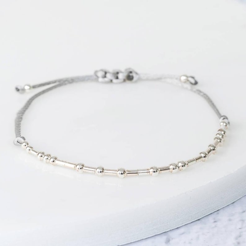 K&ouml;nnte beinhalten: Ein silbernes Perlenarmband mit einem verstellbaren grauen Band. Das Armband hat eine Reihe kleiner, runder Perlen und zylindrischer St&auml;be, die zu einem zarten Accessoire aufgereiht sind. Das Band ist zur Anpassung geknotet.
