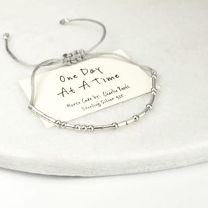 Op de afbeelding: Een zilveren Morsecode armband met de zin "One Day At A Time". De armband is geregen op een grijze koord en bevat een klein label met de naam van de ontwerper en "Sterling Silver 925". De armband wordt gepresenteerd op een wit oppervlak.