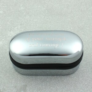May include: A silver oval-shaped box with a black rim. The box has an engraved message that reads "I'm reminded of your strength and courage!"